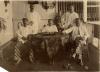 A photo of five International Drillers wearing white shirts, around a table with patterned tablecloth. There is a set of glasses on the table. There is a servant standing behind a plant against a white wall.