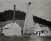 A photo of three men standing in front of an oil rig covered in wooden planks. There is a white building with a door at the front and a chimney coming out of the roof on the left. There is a line of trees in the background.