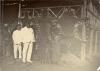 William McRae and another driller in white suits standing with a local family in front of three stretched tiger skins. 