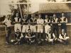 Twelve men wearing soccer uniforms in front of a net. Eight of the men are standing (seven with their arms folded) and four are sitting. There is a ball in the middle of the seated players.  