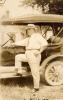 A photo of Fred Webb standing with his foot up on the step of a car. Webb is wearing a white shirt and pants, a tie, and a boat hat. The car has a canvas top and no windows. There is a building with a peaked roof in the background. 