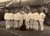 A photo of eleven International Drillers wearing white suits and holding canes. There is a man wearing dark clothes and a hat sitting in front. Behind, there is a row of buildings with triangular flags hanging between them. 