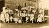 A photo of a group of men and women, with one child, in front of a thatched building. Most of the people are wearing white shirts. 