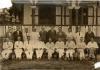 A photo of nineteen men in two rows. They are in front of a building with a deck and white railings. The back row is standing and the front row is sitting in chairs. Most of the men are wearing white.  