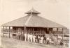 A photo of a group of men standing outside a wooden building with an ornate roof. There is a deck with a railing around the second floor with a set of stairs running up to it.