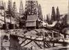 A photo of a group of wooden oil derricks. There is a narrow bridge in the foreground supporting a pipe. There is a cylindrical storage tank on the left with steam boiler stacks sticking up behind it.