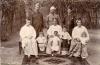 Three men stand behind three sitting women and four children. They are wearing simple shirts and skirts made from a large piece of fabric. There are trees and a wooden fence behind them, and a patterned rug at their feet.