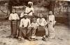 A photo of six Burmese people in traditional clothing. Three are sitting on a wicker chair, and there is a girls standing on either side. A man stands behind holding a machete.