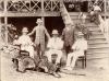 A photo of five men wearing pith helmets, two standing and three sitting in chairs, behind a large cat skin and two sets of animal horns. There are two people and a dog on a set of stairs leading up to a building behind the men.