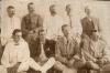 A photo of nine men. Five are sitting on a bench and four are sitting cross-legged on the floor. They are wearing a mixture of light and dark jackets.