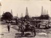 A photo of a group of men with oil derricks in the background. Some men are sitting on carts while others stand around a pile of casing. There is a set of large wagon wheels in the middle of the photo. 