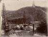 A photo of two metal oil rigs and a third rig up on a hill in behind. There are more hills in the background.