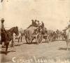 A photo of horse-drawn carts packed with goods. There are men riding on one of the carts and other men on horseback.