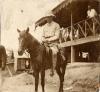 A photo of an International Driller wearing white clothing and a pith helmet, sitting on a horse. He is in front of a building that has been raised on stilts.