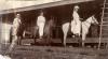 Deux hommes et une femme à cheval devant un bâtiment avec une terrasse surélevée. Les hommes montent des chevaux foncés et portent des chemises et des pantalons blancs. La femme est vêtue d'une robe blanche et d'un chapeau. Elle monte un cheval blanc. 