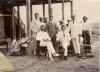 Five International Drillers pose under the stilts which support a house. They are wearing light-coloured suits. Seven local people stand behind them.