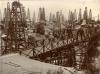 A field full of four-poled oil derricks behind a bridge spanning a low point in the field. There are buildings behind the first row of derricks.