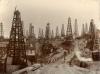 Close to thirty oil derricks with four poles each. There is a bridge leading across a low point in the oil field and several men are walking along the road. There are buildings behind the front row of derricks and a large groups of people are gathered near a pile of casing.