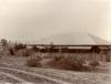 A bungalow with thatched window covers. It is surrounded by a fence made of sticks. There is a cart parked by the fence and a cluster of shrubs in front of it. There is a group of oil derricks in the background.