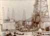 An oil crew in front of an oil rig. There are five oil rigs in the background and a pile of casing at the front of the photo. An International Driller is sitting on some of the casing.