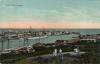 A postcard showing the entrance to a harbour.. There are boats docked along the edge of the light blue water. A group of people is standing on a hill looking at the boats. There are buildings very close to the edge of the water.  