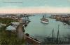 A postcard depicting a small bay with light blue water. There is a boat with two masts sailing in the water and a number of boats tied along docks. There are buildings close to the edge of the water. The postcard says "Babia de Santa Ana, Curacao". 