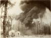 A photo of a column of dark smoke behind two buildings. There is a group of oil derricks on the right.  