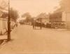  A dirt road with buildings and fences on either side. There are people walking on the street and there is a covered cart, as well. 