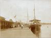 A ship docked alongside a wooden dock. There is another ship in behind and a building to the left. People are sitting and standing, looking at the ships. 