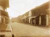 A dirt street with curbs and buildings on both sides which are close together. At the far end of the street a group of people is walking close together. 