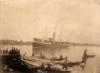 A ship with two masts and a smoke stack floating in the water. There are buildings and trees on the land behind it. In the foreground there is a group of people at a dock with a number of small boats. 