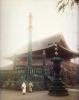 A photo of two Japanese people in traditional clothing, standing in front of a temple. There are cherry blossoms in bloom behind a stone wall and to the right of the image. The photo has been tinted with pinks, yellows, blues, and greens.  