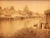 A photo of a group of buildings on stilts with thatched roofs. They are sitting directly above water and there is a bridge between the two sides of the river towards the back. There are trees in the background and reeds in the foreground.  