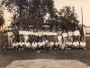 A baseball team. Half the men are standing and half are sitting. Most are wearing white t-shirts. They are in front of a group of trees and a building. There are three baseball bats set up like a tripod in front of them with a ball resting on top. Some other equipment is also in front of them. 