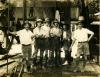 A photo of six International Drillers standing inside an oil rig with metal siding. They are wearing shorts, high socks, and white t-shirts.