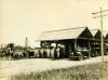 A group of people standing in front of an open wooden structure with a metal roof that has three peaks. There is a car and horse on either side of the group.