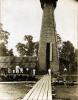 A photo of W.B.C. Isbister in a white suit and his dog standing on a plank path in front of a thatched oil rig. His crew stands to the left on a pile of casing.