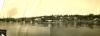 A panoramic photo of a buildings at the edge of a waterway. There are boats on the water in the foreground and oil rigs along the hills in the background.