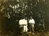 Une photo de trois hommes vêtus de blanc, debout dans la jungle. L'homme de gauche est détourné et porte un chapeau. 