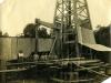 A photo of eight men standing behind a pile of casing and in front of an oil rig that is covered in metal sheeting.