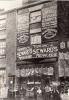 A brick building with birdcages hanging from the top of the windows and along ropes strung across to the next building. There is a group of people outside. Signs on the store front advertise "Trading - Seward's Menagerie" and the animals they sell. 