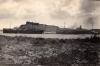 A photo of two ships tied to a dock on a pier. There is a hill in the background and gravel and plants in the foreground. 