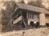 A photo of a bungalow with a thatched roof and sides built on stilts. A dog and a woman sit on the steps going up to the deck, and a man stands to the side of the steps. 