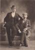 Charles Wallen leaning on a chair that Alf Burns is sitting in. They are wearing dark suits and hats, and are holding pipes. The background is a dark wall. 