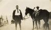 George Rawlings dans un complet foncé et des bottes d’équitation. Il tient un casque colonial dans une main et les reines du cheval dans l’autre. Un homme se tient derrière le cheval.  