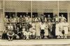 A group of International Drillers and their families outside of a building with latticework on the porch. The men are wearing suits and the women are wearing dresses. 