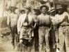 A group of men in hats and dirty pants. Nine are standing and one is sitting. The man third from the left is wearing a light suit and tie. 