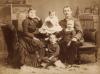 A woman and man sitting with three children between them. They are wearing dark clothing except for the youngest child in the middle who is wearing white. There is a mantle behind them and a fur rug at their feet. 