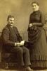 A photo of Charles and Jessie Nicklos. Charles is sitting in a chair and holding a piece of paper while Jessie is standing with her arms folded. 