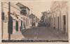 The front of a postcard showing a street with cars and a team of horses in the road.